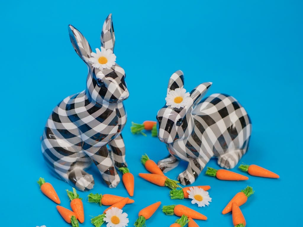 Playful checkered bunny figurines surrounded by carrots and flowers on a vibrant blue background, perfect for Easter celebrations.